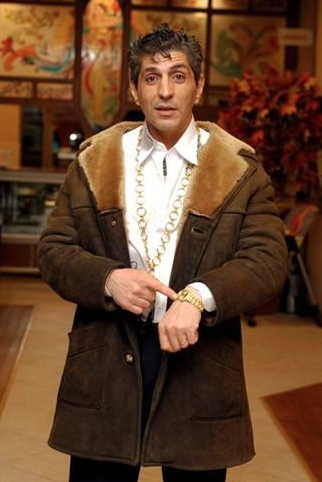 Erkan Aydoğan Oflu im Ledermantel mit Pelzkragen, weissem Hemd und viel Gold um den Hals. Er zeigt auf eine Uhr.