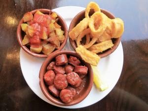 Dreierlei spanische Tapas. Kartoffelecken mit Tomatendip, frittierte Tintenfischringe und Chorizo
