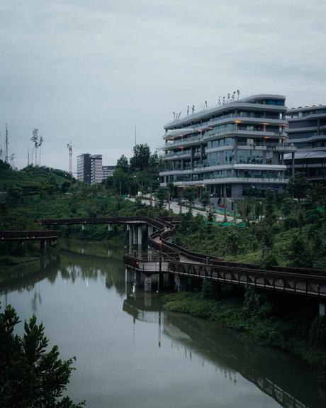 Nusantara 2025: Unsere Reise in Indonesiens neue Hauptstadt Das moderne Gebäude von Nusantara steht anmutig neben einem gewundenen Fluss und einer Fußgängerbrücke, eingebettet in üppiges Grün unter einem wolkigen Himmel.