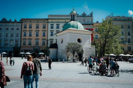 Das perfekte Krakau-Wochenende Menschen gehen spazieren und versammeln sich um eine kleine weiße Kapelle auf einem belebten Stadtplatz mit farbenfrohen Gebäuden dahinter.