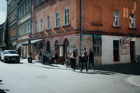 Das perfekte Krakau-Wochenende Menschen gehen spazieren und sitzen vor einem Café an einer sonnigen Straßenecke in der Stadt mit orangefarbenen und beigen Gebäuden.