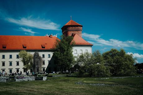 Das perfekte Krakau-Wochenende Ein historisches Gebäude mit roten Dächern, Bäumen und Menschen, die unter blauem Himmel auf einer Rasenfläche spazieren.