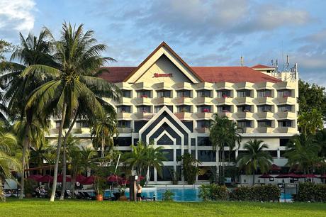 Digitaler Nomade in Tansania: Arbeiten zwischen Serengeti und Sansibar Das Miri Marriott Resort ist ein großes Hotel mit einem markanten roten Dach und mehreren Balkonen, ergänzt durch einen glitzernden Swimmingpool, der von Palmen umgeben ist. Der Himmel ist teilweise bewölkt und wirft sanfte Schatten auf den Rasen im Vordergrund.