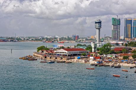 Digitaler Nomade in Tansania: Arbeiten zwischen Serengeti und Sansibar Stadtbild am Wasser mit Booten, Menschenmassen und modernen Gebäuden unter einem bewölkten Himmel.