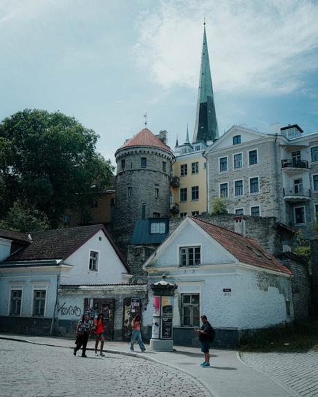 Tallinn: Die 10 besten Sehenswürdigkeiten Unter einem teilweise bewölkten Himmel gehen Menschen in Tallinn an alten Steingebäuden und einem Turm mit spitzer Spitze vorbei.