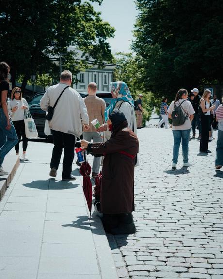 Tallinn: Die 10 besten Sehenswürdigkeiten Eine Frau in einem braunen Mantel kniet auf einer Kopfsteinpflasterstraße in Tallinn und hält eine Tasse in der Hand, während Menschen vorbeigehen.