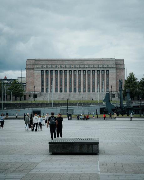 Helsinki: Die 10 besten Sehenswürdigkeiten Menschen gehen auf einem großen Platz in Helsinki spazieren, mit dem finnischen Parlamentsgebäude und einer dunklen Skulptur im Hintergrund.
