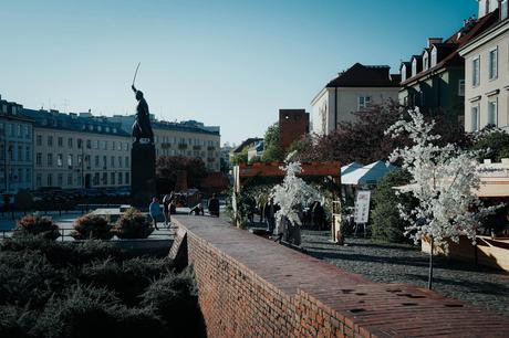 Helsinki: Die 10 besten Sehenswürdigkeiten An einem sonnigen Tag gehen Menschen auf einem europäischen Stadtplatz an einer Statue und Marktständen entlang einer Backsteinmauer vorbei.