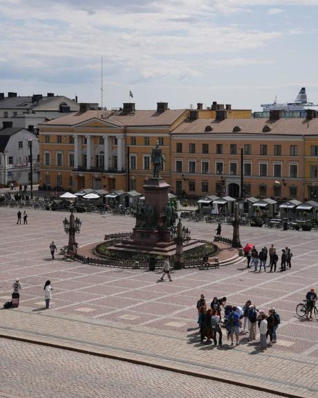 Helsinki: Die 10 besten Sehenswürdigkeiten Menschen versammeln sich um eine große Statue in der Mitte eines kopfsteingepflasterten Platzes in Helsinki, mit gelben Gebäuden im Hintergrund.