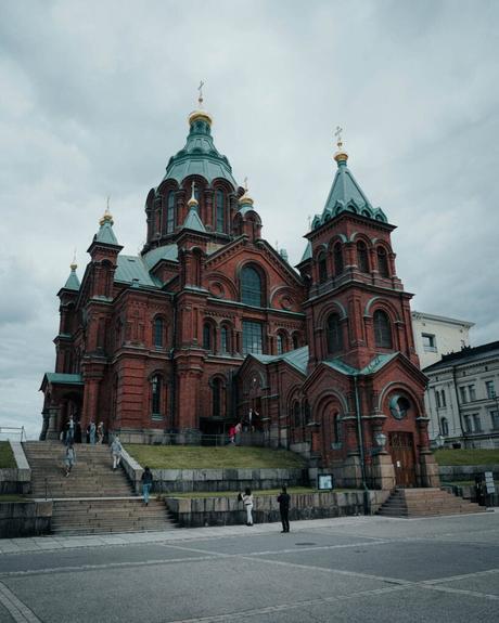Helsinki: Die 10 besten Sehenswürdigkeiten Eine Kathedrale aus rotem Backstein mit grünen Kuppeln und goldenen Kreuzen steht unter dem wolkenverhangenen Himmel Helsinkis, Menschen gehen in der Nähe spazieren.