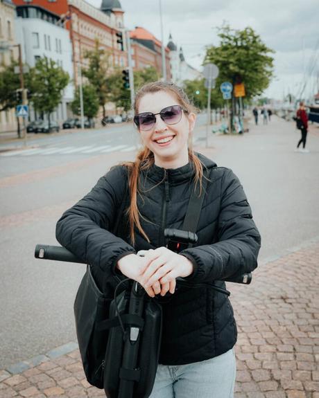 Helsinki: Die 10 besten Sehenswürdigkeiten Eine lächelnde Frau mit Sonnenbrille und schwarzer Jacke steht auf einer Straße in Helsinki und ruht sich auf einem Elektroroller aus.