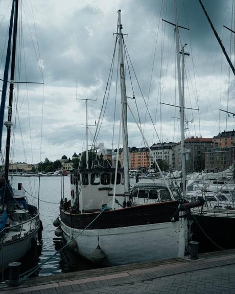 Helsinki: Die 10 besten Sehenswürdigkeiten Ein angedocktes Segelboot mit Masten unter einem wolkenverhangenen Himmel in Helsinki, vor einem Hintergrund aus bunten Gebäuden.