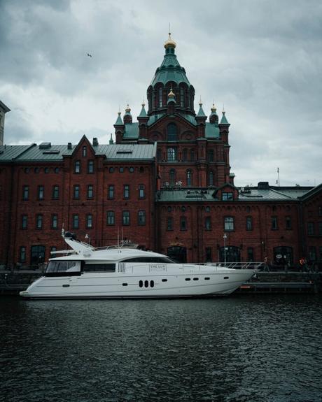 Helsinki: Die 10 besten Sehenswürdigkeiten Eine weiße Yacht, die an einem roten Backsteingebäude mit grünen Kuppeln angedockt ist, unter einem bewölkten Himmel in Helsinki.