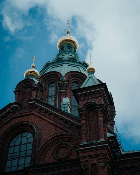 Helsinki: Die 10 besten Sehenswürdigkeiten Kirche aus rotem Backstein mit grünen Kuppeln und goldenen Kreuzen vor einem wolkenverhangenen blauen Himmel über Helsinki, von unten gesehen.