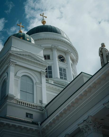 Helsinki: Die 10 besten Sehenswürdigkeiten Weiße Kirche mit grünen Kuppeln, goldenen Kreuzen und einer Uhr vor einem teilweise bewölkten Himmel in Helsinki.
