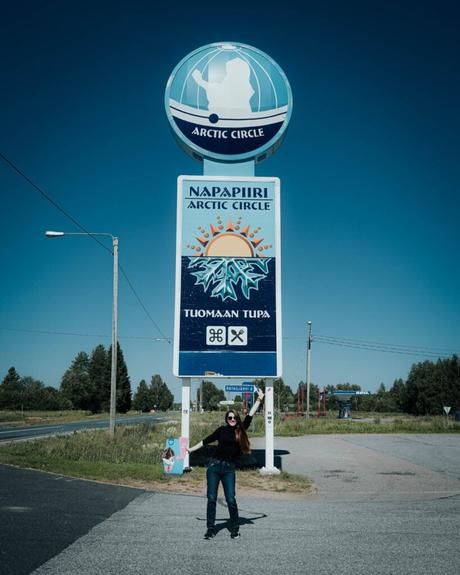 Im Bann des Mitternachtslichts: Ein Abenteuer in Finnisch-Lappland Eine Frau steht lächelnd unter einem großen Polarkreisschild am Straßenrand, umgeben von Grün und blauem Himmel.