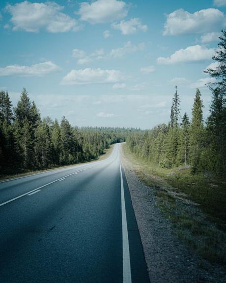 Im Bann des Mitternachtslichts: Ein Abenteuer in Finnisch-Lappland Eine lange, leere Straße erstreckt sich in der Ferne, gesäumt von Kiefern unter einem blauen Himmel mit vereinzelten Wolken.