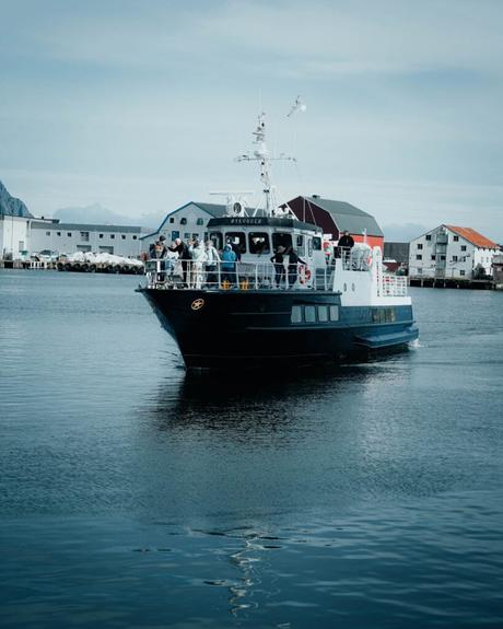 Svolvær und die Teufelspforte Ein kleines Passagierboot mit Menschen an Bord fährt an einem ruhigen, bewölkten Tag in der Nähe der Hafengebäude.