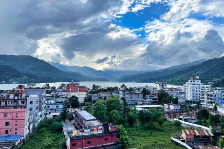 Svolvær und die Teufelspforte Bunte Gebäude und üppiges Grün umgeben einen glitzernden See in Pokhara, dessen Berge sich unter einem dramatischen, wolkenverhangenen Himmel erheben.