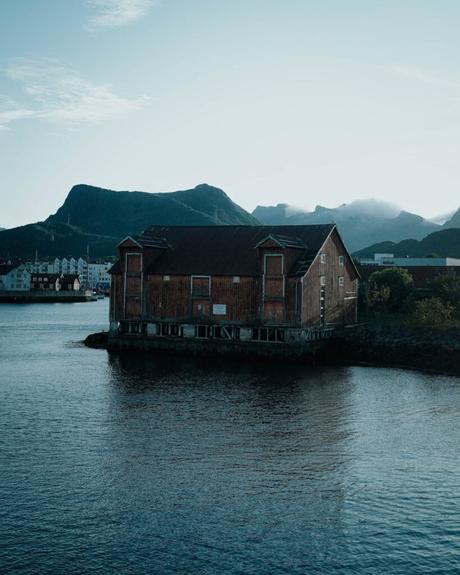 Svolvær und die Teufelspforte Ein verwittertes Holzgebäude steht am Ufer des Wassers, im Hintergrund sind die Berge zu sehen.