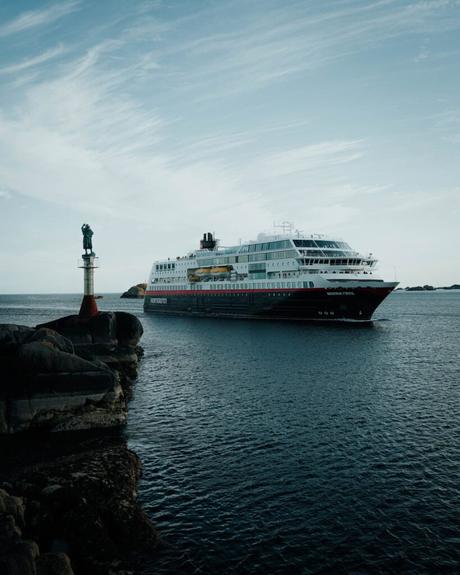 Svolvær und die Teufelspforte Ein Kreuzfahrtschiff fährt an einem Leuchtturm mit einer Statue vorbei, in der Nähe des felsigen Ufers unter einem teilweise bewölkten Himmel.