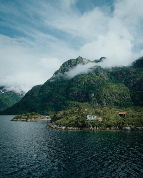 Svolvær und die Teufelspforte Üppig grüne Berge mit Wolken, Hütten am Seeufer und ruhiges Wasser unter einem teilweise bewölkten Himmel.