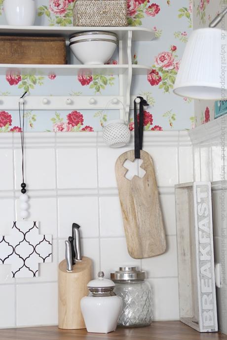 Blick in eine Küchenecke mit geblümter Tapete, Wandregal, und Holzfarbenen sowie weißen Utensilien und einer Cross-Chain