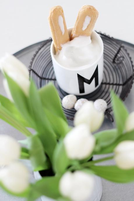 Design Letters Tasse mit Löffelbiskuits als Hasenohren, daneben stehen weiße Tulpen