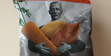 Schwarzweiß Bild eines Mannes der farbliches, überdimensional großes Gemüse in der Hand hält auf der Tyrrells Veg Crisps Packung 