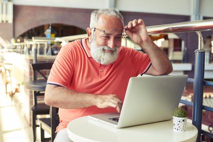 cheerful senior man raised his glasses and looking at laptop in cafe