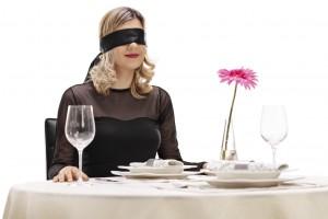Young woman with blindfold on her eyes sitting at a romantic dinner table isolated on white background