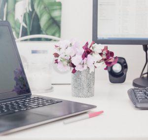 Ein Schreibtisch, auf dem zwischen Laptop und Monitor ein kleiner Strauß Blumen in einer Vase steht