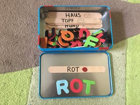 Metalldose mit bunten magnetischen Buchstaben und Holzspateln. Auf den Holzspateln sind einfache Wörter aufgeschrieben und ein passendes Bild aufgemalt. Aufgabe ist es die Wörter mit den magnetischen Buchstaben nachzulegen. Dieses Spiel ist einer der ersten Schritte zum Lesen lernen.
