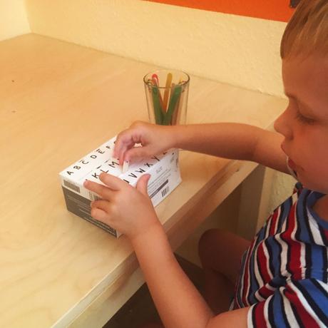 Kind spielt mit Buchstaben Steckspiel. Die Buchstaben sind auf Eissteilen geschrieben, die in den passend beschrifteten Schlitz in einer Pappschachtel gesteckt werden müssen. Dieses Spiel ist einer der ersten Schritte zum Buchstaben und schließlich zum Lesen lernen.