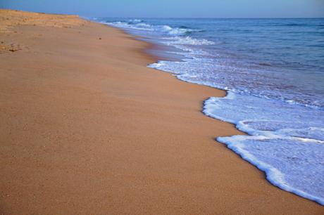 Wassertemperatur Faro: Orangefarbener Sandstrand mit weiß schäumenden, auslaufenden Wellen
