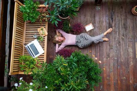 Seniorin entspannt auf eigener Terrasse mit Essen, Laptop und wirkt gelassen inmitten von Pflanzen.