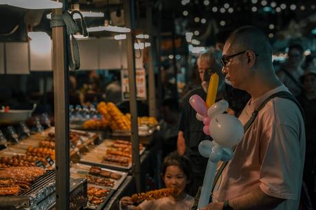 Ein Mann mit Ballontieren steht neben einem Kind, das sich an einem Spieß gütlich tut, während sie gemeinsam den lebhaften Nachtmarkt von Fotospots Bangkok erkunden, mit Essensständen voller aufgespießter Köstlichkeiten, alle in warmes Deckenlicht getaucht.