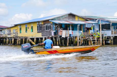 Person, die mit einem kleinen Motorboot an bunten Pfahlhäusern auf einem Fluss vorbeifährt.