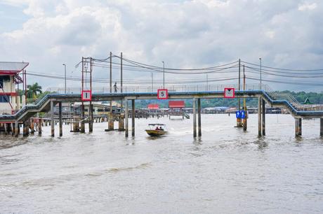 An einem bewölkten Tag fährt ein kleines Boot unter einer Fußgängerbrücke hindurch, im Hintergrund sind Gebäude am Wasser und Bäume zu sehen.