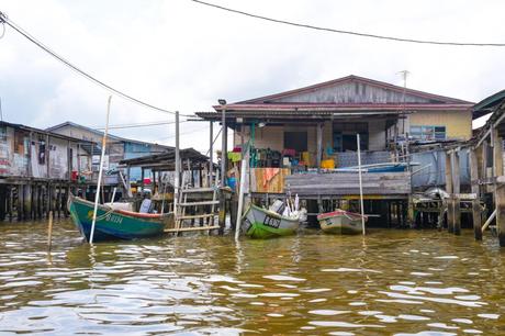 Holzhaus auf Stelzen am Wasser mit kleinen Booten, die in der Nähe angedockt sind. Bewölkter Himmel im Hintergrund.