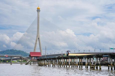 Hohe Hängebrücke über einem Fluss mit zwei Personen, die auf einem Holzpfeiler laufen, vor einem bewölkten Himmel.
