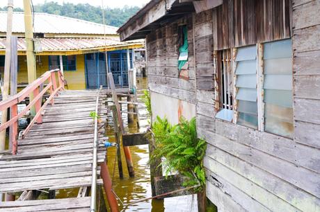 Holzhäuser auf Pfahlbauten mit einem alten Gehweg über dem Wasser, umgeben von Grün und anderen ähnlichen Bauten in der Nähe.