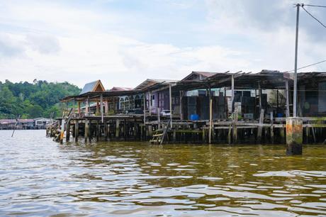 Pfahlhäuser über dem Wasser mit üppigen grünen Hügeln im Hintergrund unter einem teilweise bewölkten Himmel.