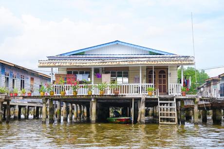 Holzpfahlhaus über dem Wasser mit Pflanzen und Wäsche, die auf der Veranda hängt. Darunter liegt ein kleines Boot.