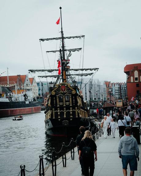 Ein großes Piratenschiff liegt an einer belebten Uferpromenade mit Menschen und farbenfrohen Gebäuden in der Nähe.