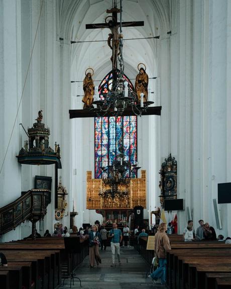 Menschen gehen und sitzen in einer großen Kirche mit hohen Decken und bunten Buntglasfenstern.