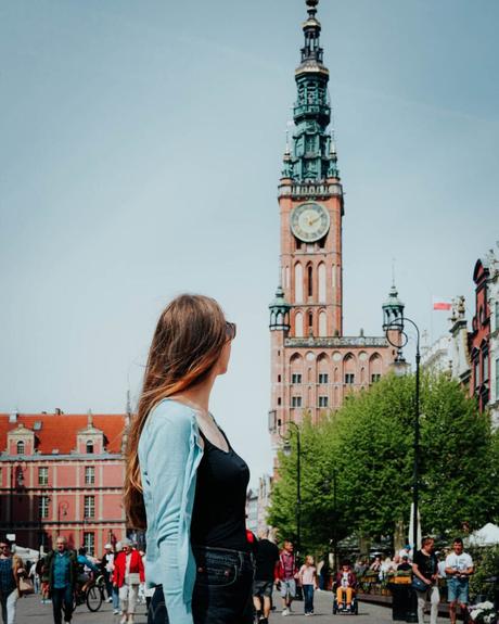 Eine Frau mit langen Haaren steht auf einem belebten Stadtplatz und blickt auf einen hohen Uhrenturm.
