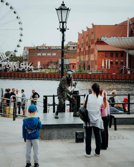 Ein als Statue verkleideter Straßenkünstler unterhält die Menschen in der Nähe eines Flusses mit einem Riesenrad und roten Backsteingebäuden.