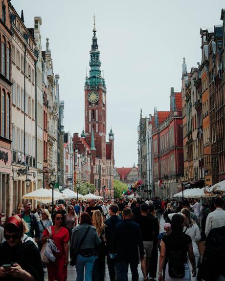 Überfüllter Straßenmarkt mit farbenfrohen Gebäuden und einem hohen Uhrenturm im Hintergrund unter einem bewölkten Himmel.
