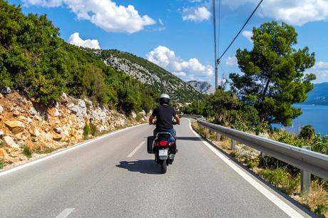 Person, die auf einem Motorroller auf einer kurvenreichen Küstenstraße mit Bergen und blauem Himmel im Hintergrund fährt.